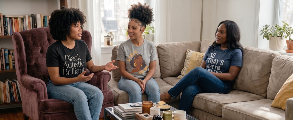 Three women sitting and conversing in a bright living room. The woman on the left wears a black graphic t-shirt that reads 'Black Autistic Brilliant!' and blue jeans, seated in a purple armchair. The woman in the middle is wearing a grey t-shirt with a graphic of a Black woman and says "Be Kind To Your Mind." The Woman on the right is wearing a dark blue t-shirt with the words "So THATS why I'm Like This."
